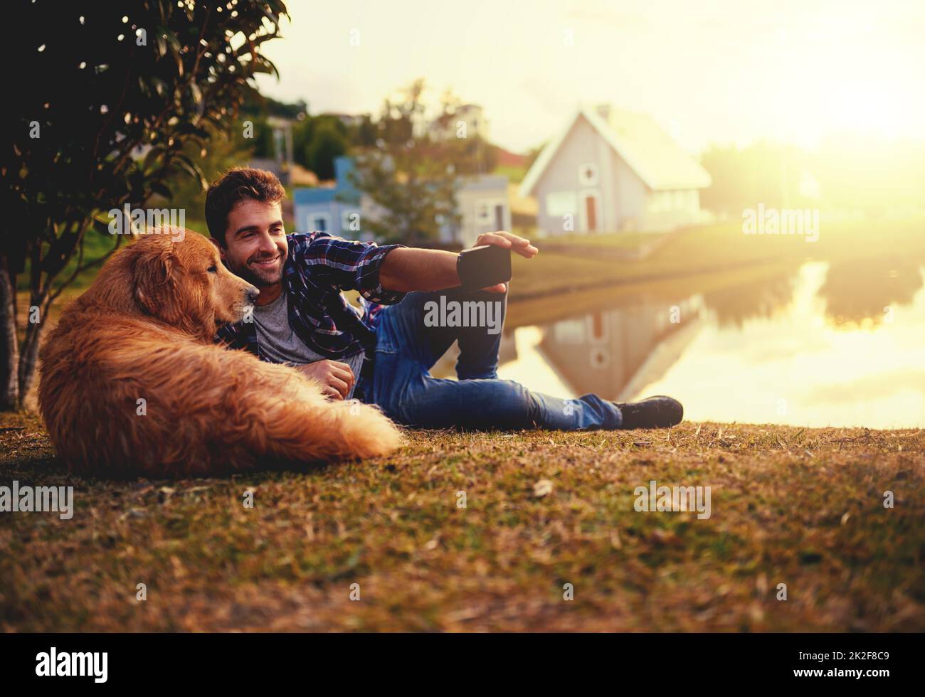 Just one more then we can head home. Full length shot of a handsome young man and his dog taking selfies by a lake in the park. Stock Photo