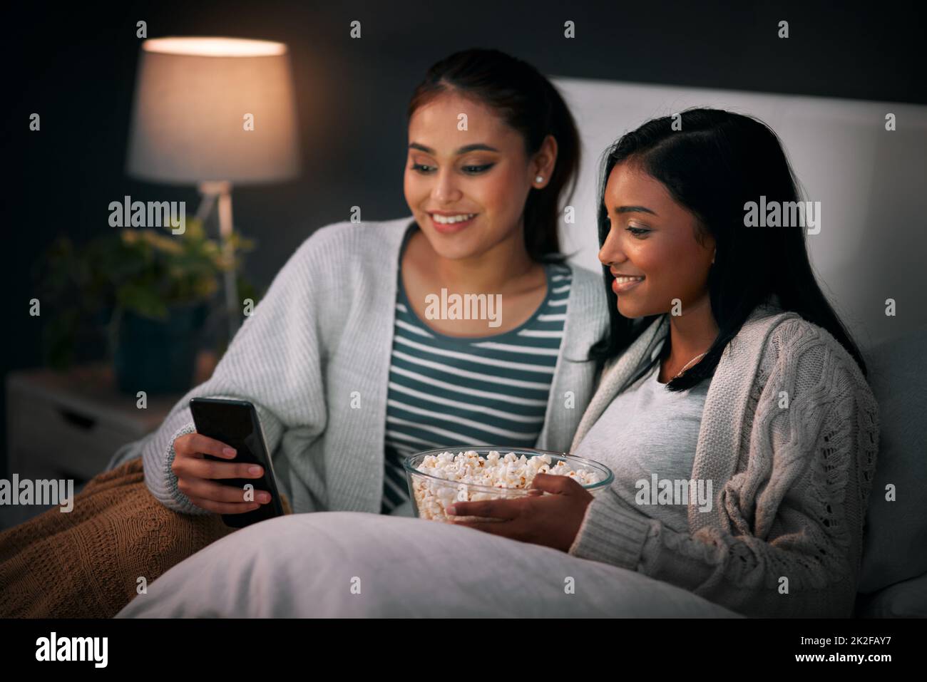 Should I invite her over. Shot of a young woman showing her friend something on her cellphone while sitting together on a bed. Stock Photo