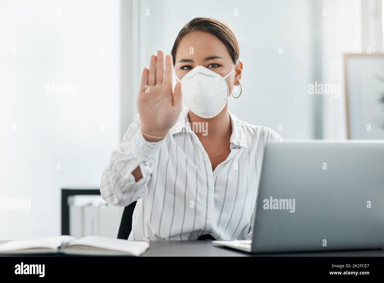 Keep your distance for your safety and mind. Shot of a masked young businesswoman gesturing to stay away while working at her desk in a modern office. Stock Photo