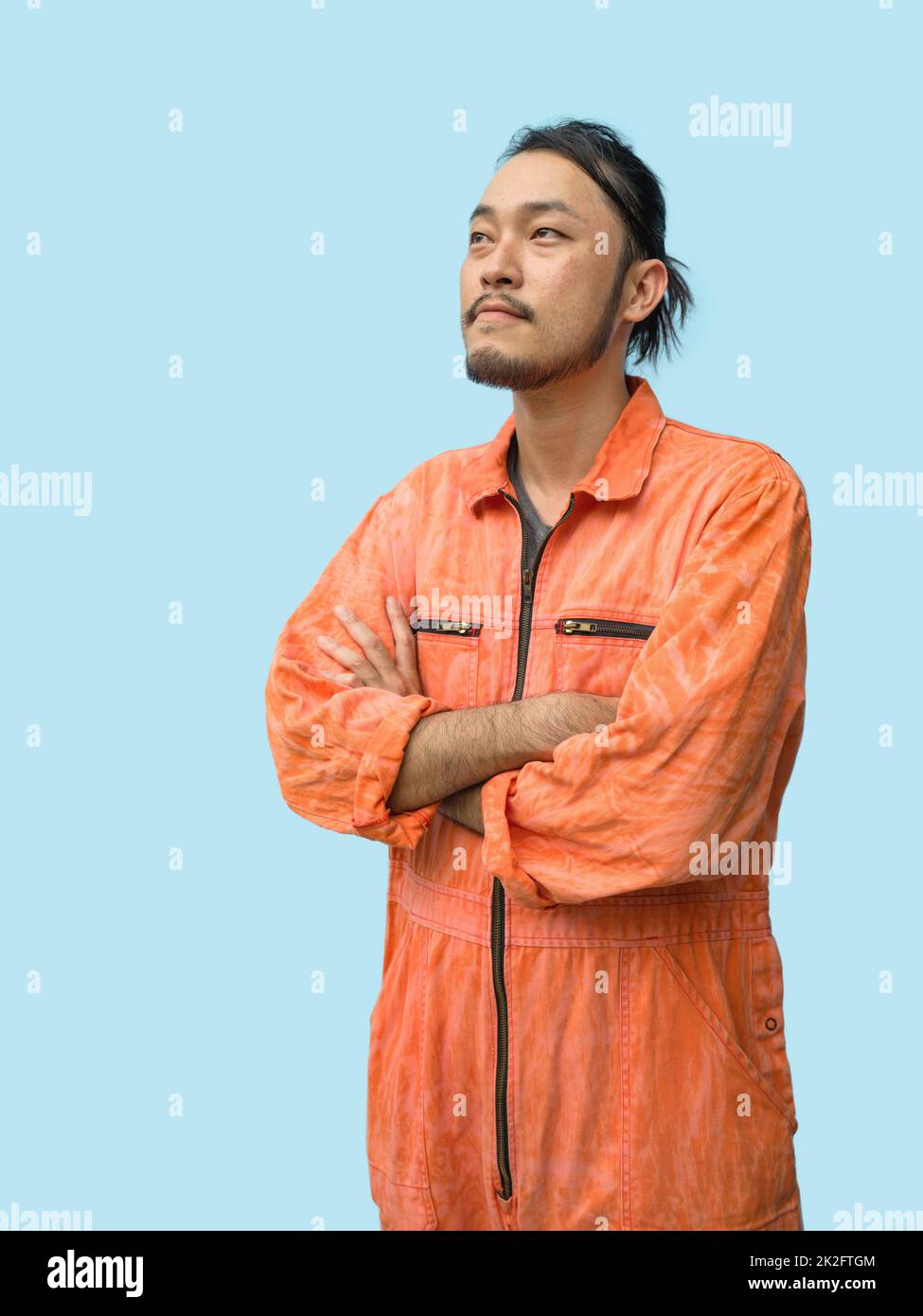 Young long hair asian man with mustache and beard stand with his arms crossed. He looked outside while thinking of something.  Portrait of industrial people on blue background. Stock Photo