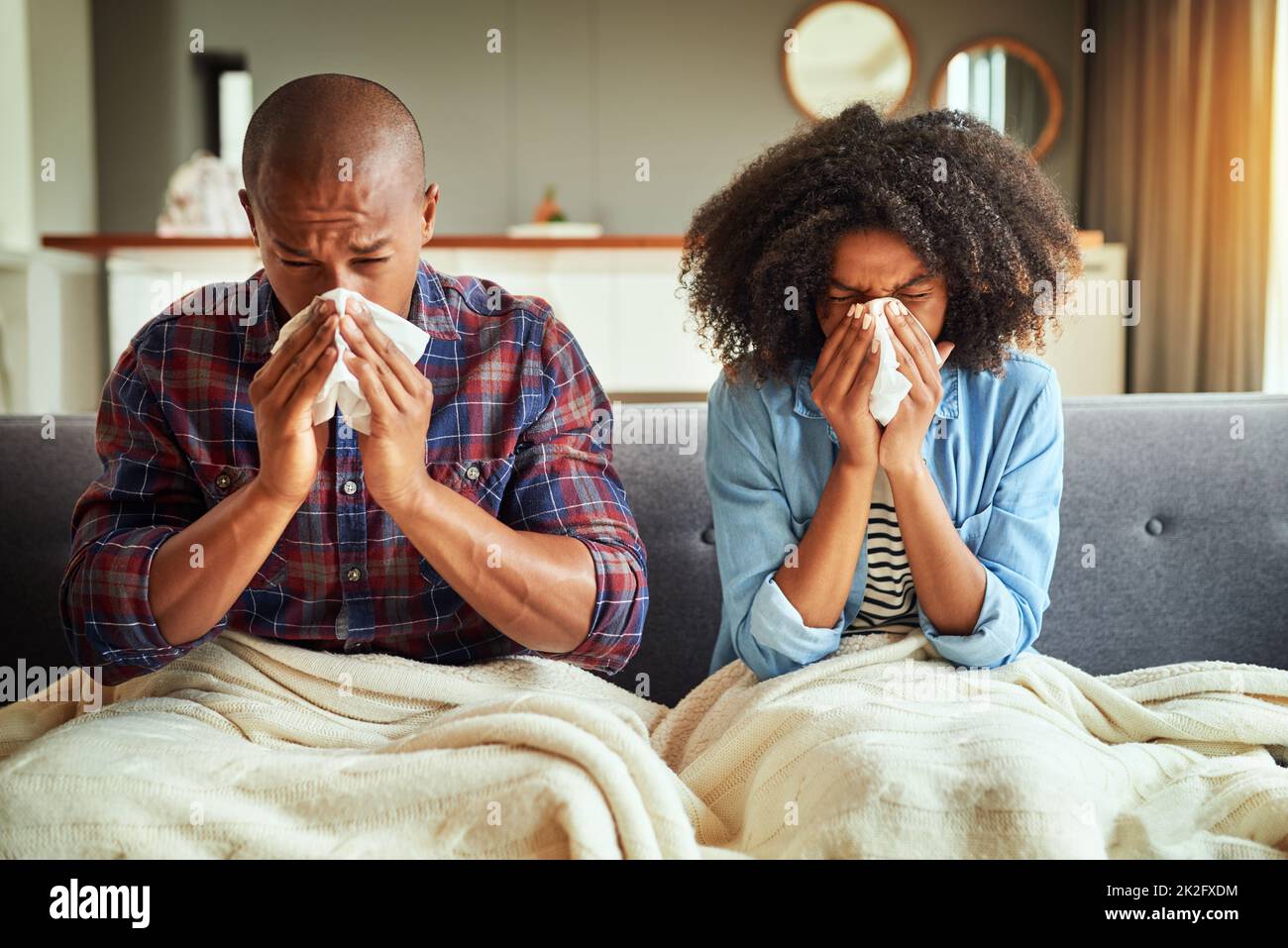 How did we both get sick. Shot of a tired looking young couple seated on a couch with blankets while being sick together at home. Stock Photo