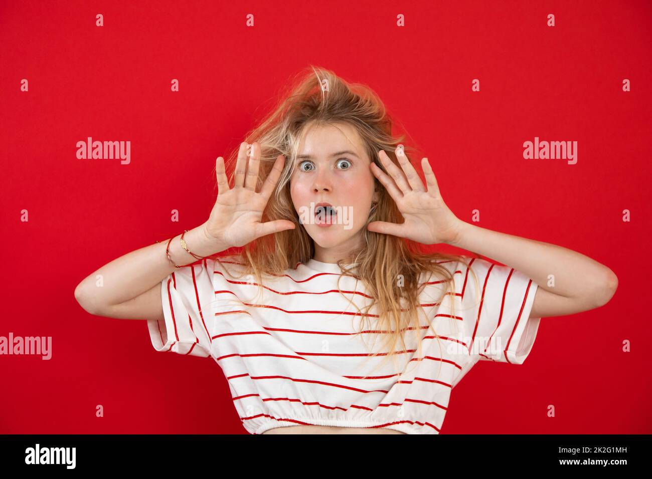 Open eyed young lady stand with raised hands with spread fingers near face on empty red background. Nice girl with wild hair have fun and show Stock Photo