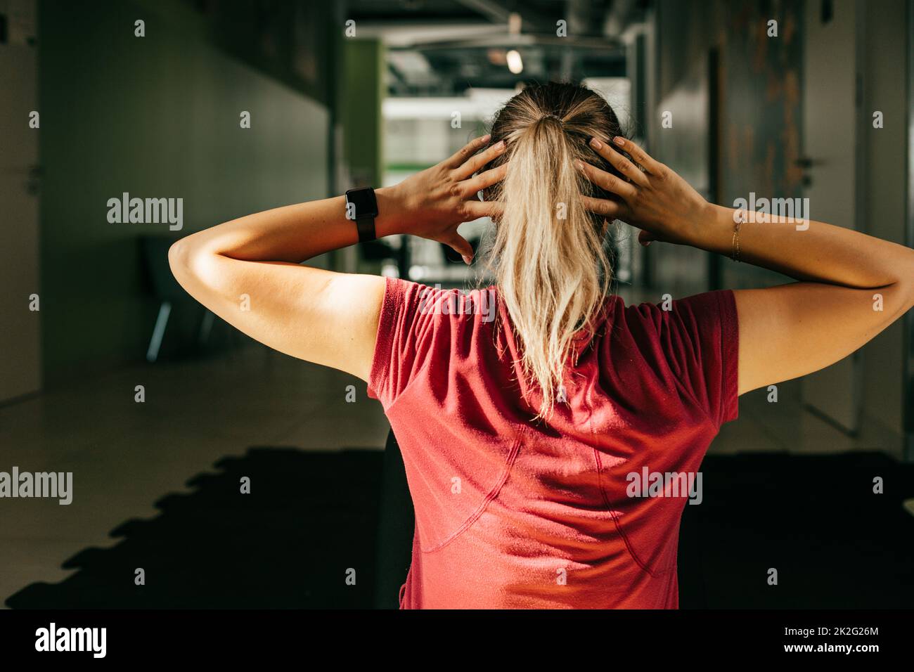 Girl with hair tied in ponytail pump press in fitness gym keeping hands behind head, back view. Plus size woman do cardio workout by lifting body Stock Photo