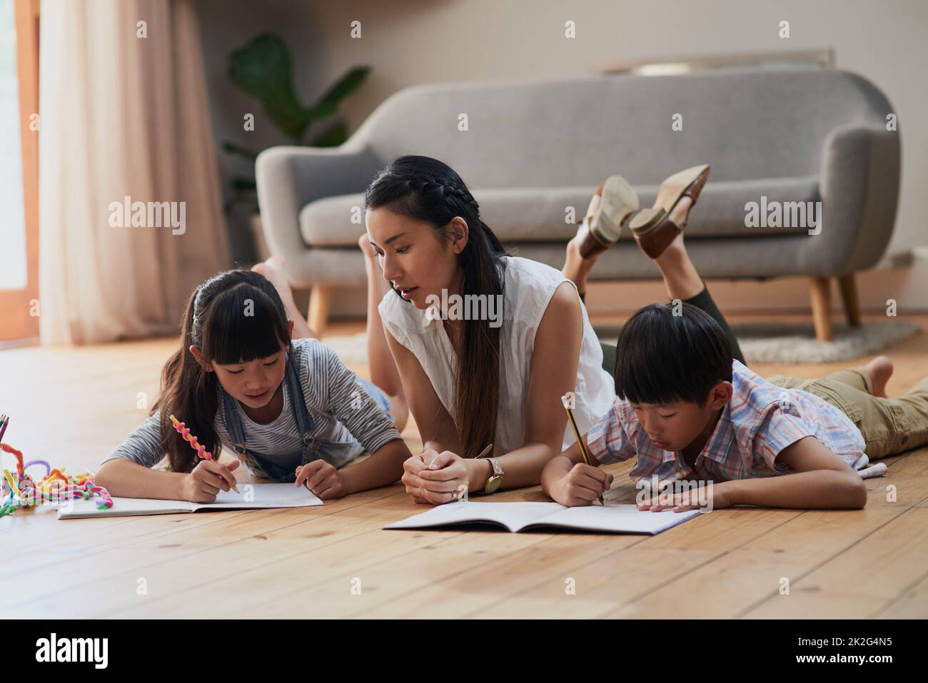 Mom makes sure that the kids do homework. Shot of a cheerful mother and her two children doing homework together while lying on the floor at home during the day. Stock Photo