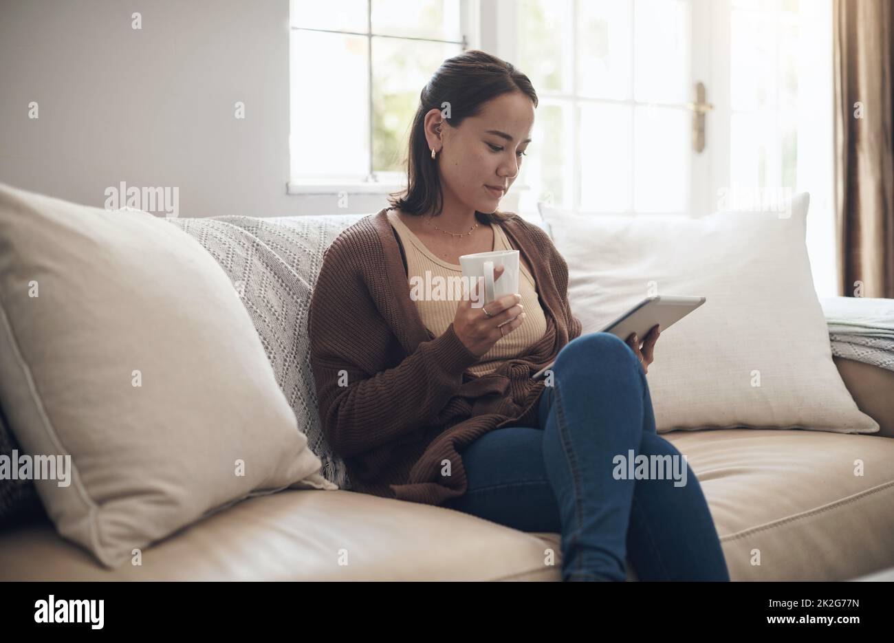 Coffee and a good ebook pairs so well. Shot of a young woman using a digital tablet while drinking coffee at home. Stock Photo