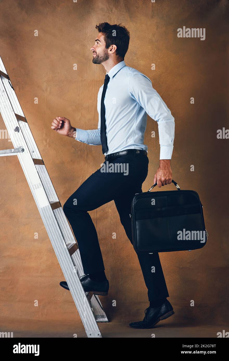 Up is the only option. Studio shot of a handsome young businessman climbing a ladder against a brown background. Stock Photo