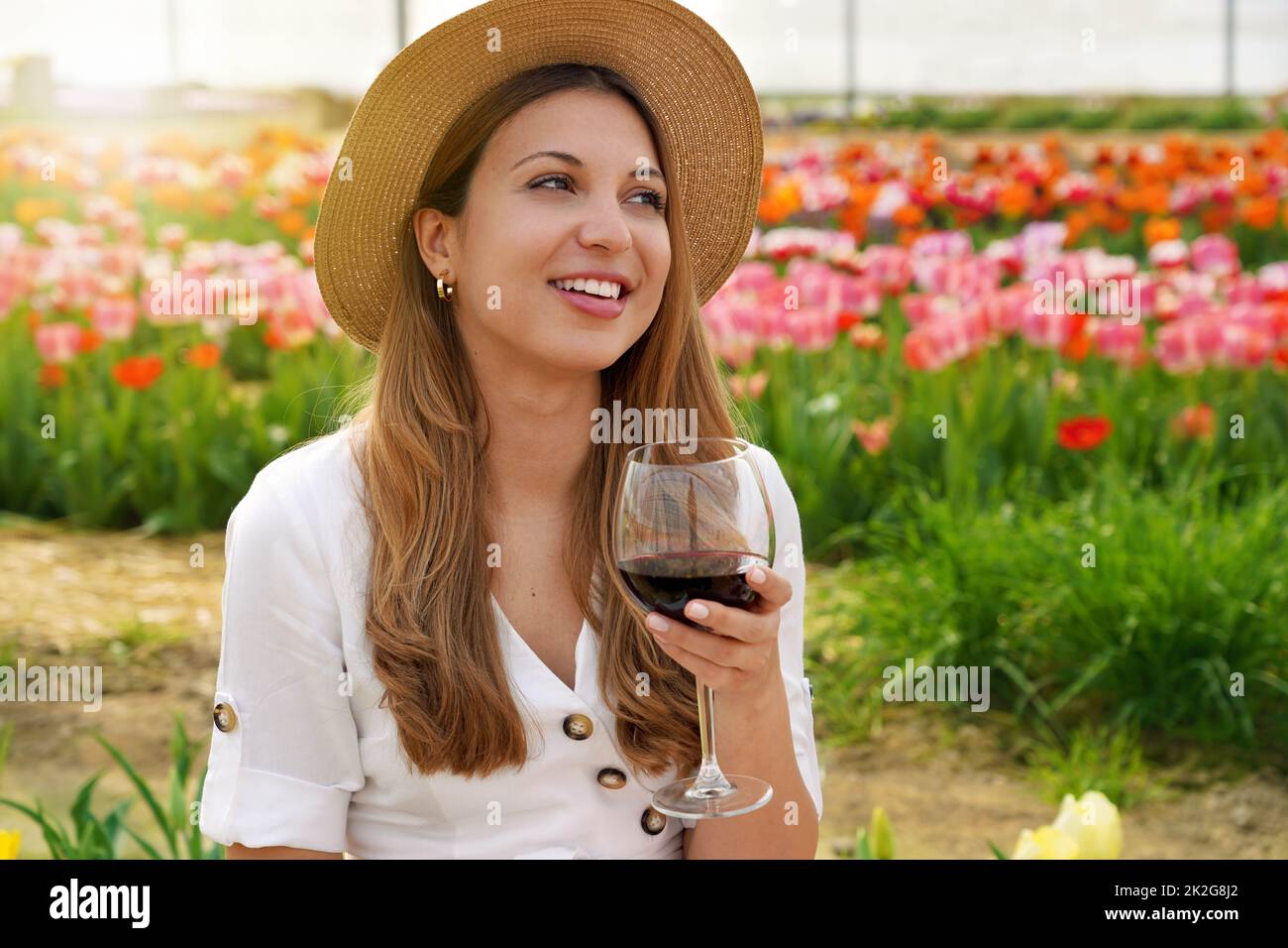 Young beautiful woman with straw hat drinking glass of red wine over flowered background looking to the side with happy face smiling Stock Photo