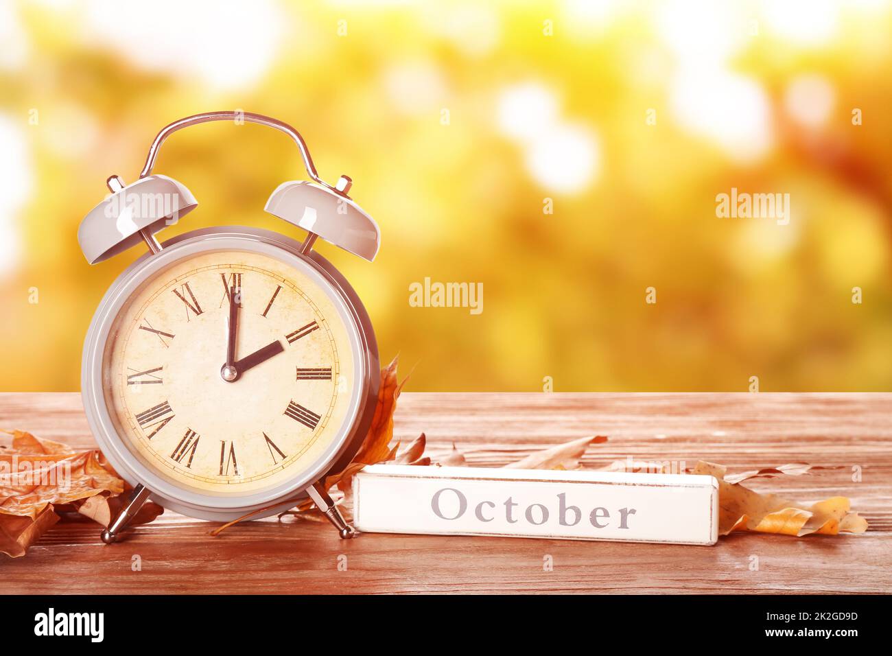 Alarm clock, word OCTOBER and autumn leaves on table outdoors. Daylight saving time end Stock