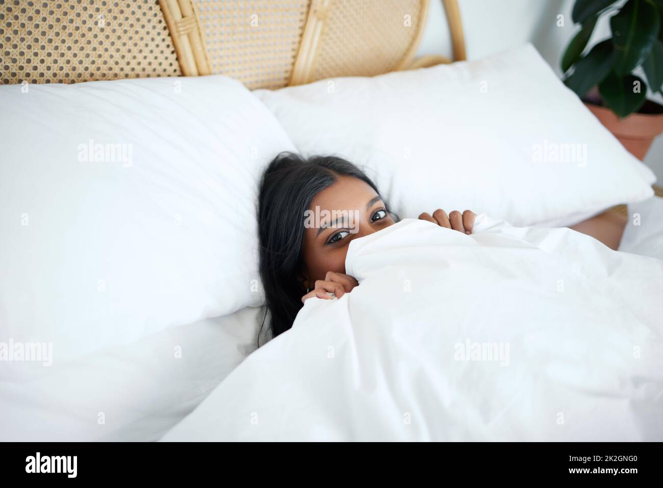 Dont tell anyone that Im here. Shot of a young woman peeking from under her duvet while lying in her bed. Stock Photo