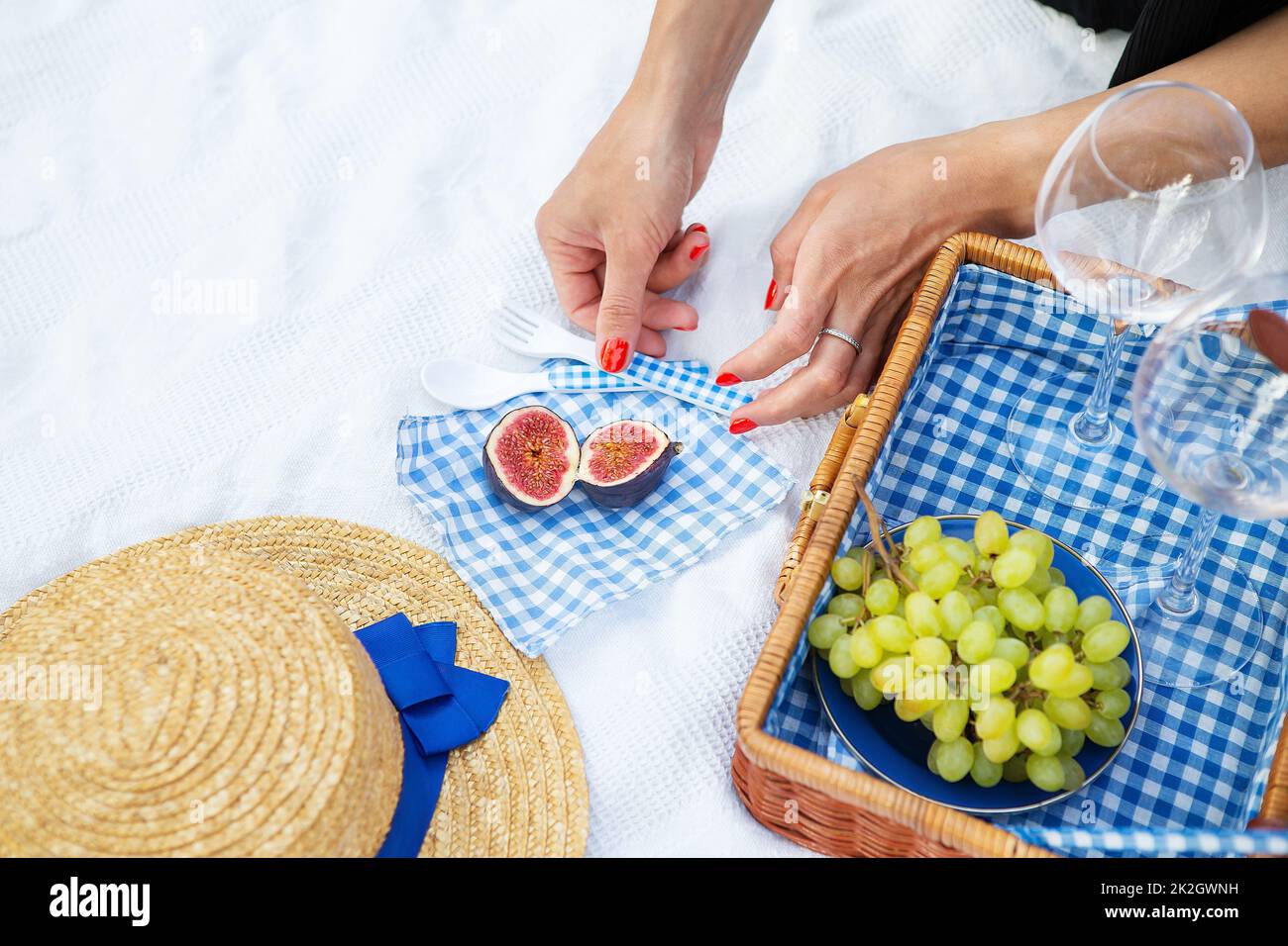 Romantic picnic in the park on the grass, delicious food: basket, wine, grapes, figs, cheese, blue checkered tablecloth, two glasses of wine. Girl cuts figs.Outdoor recreation concept. Stock Photo