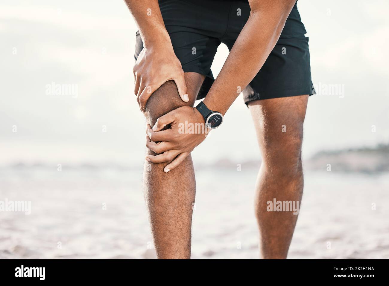 Looks like no more running. Cropped shot of an unrecognizable male athlete holding his knee in pain while exercising on the beach. Stock Photo