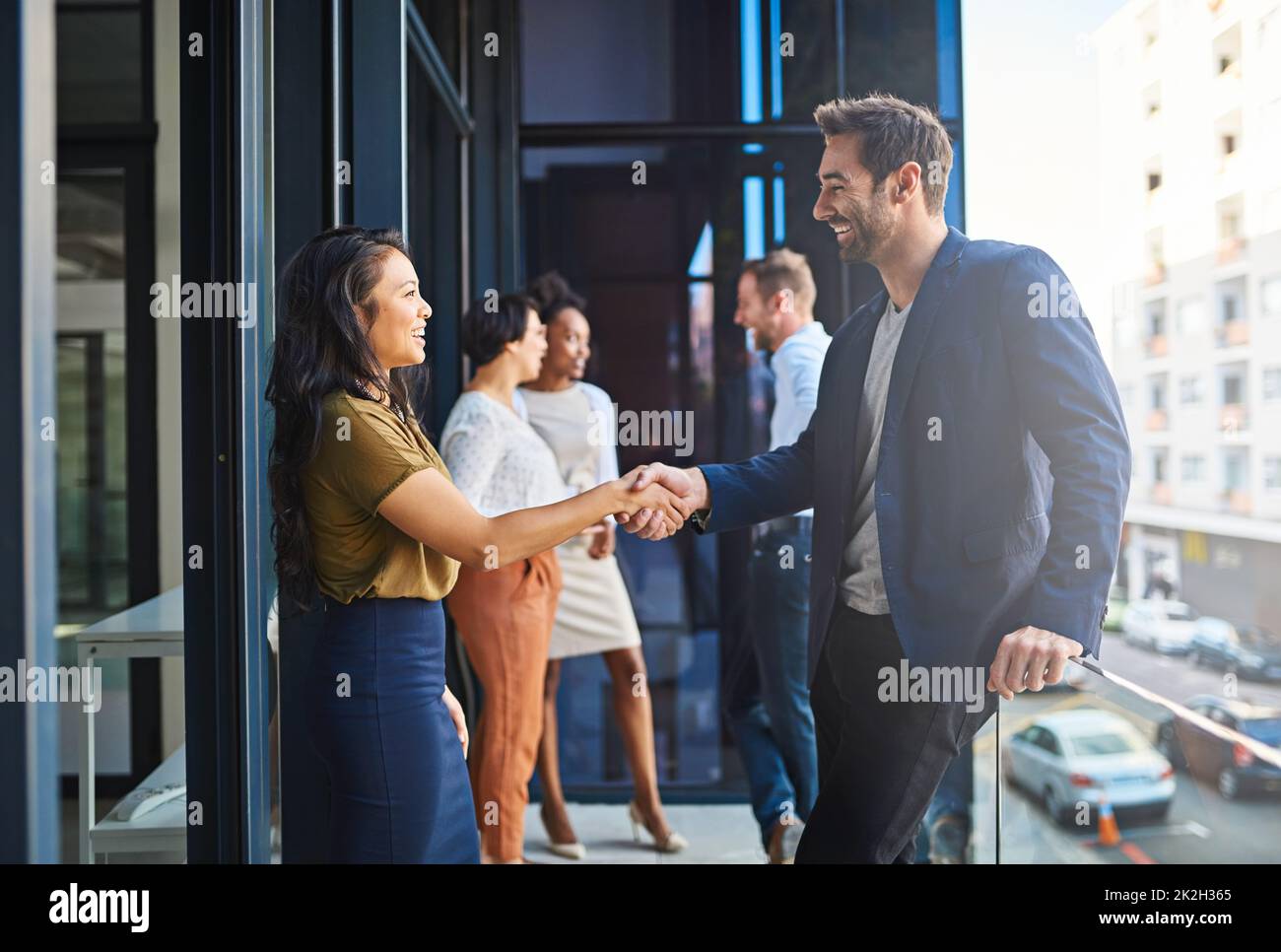 We cant wait to work with you. Cropped shot of two businesspeople shaking hands on a balcony with their coworkers in the background. Stock Photo