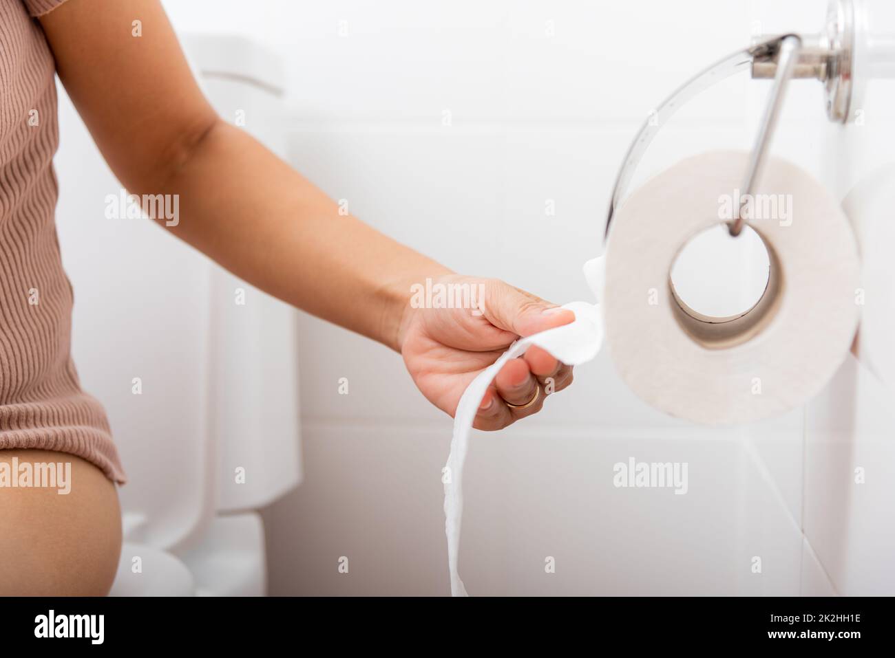 Closeup hand pulling toilet paper roll in holder for wipe.Closeup hand pulling toilet paper roll in holder for wipe, woman sitting on toilet she taking and tearing white tissue on wall to towel clean in bathroom, Healthcare concept Stock Photo