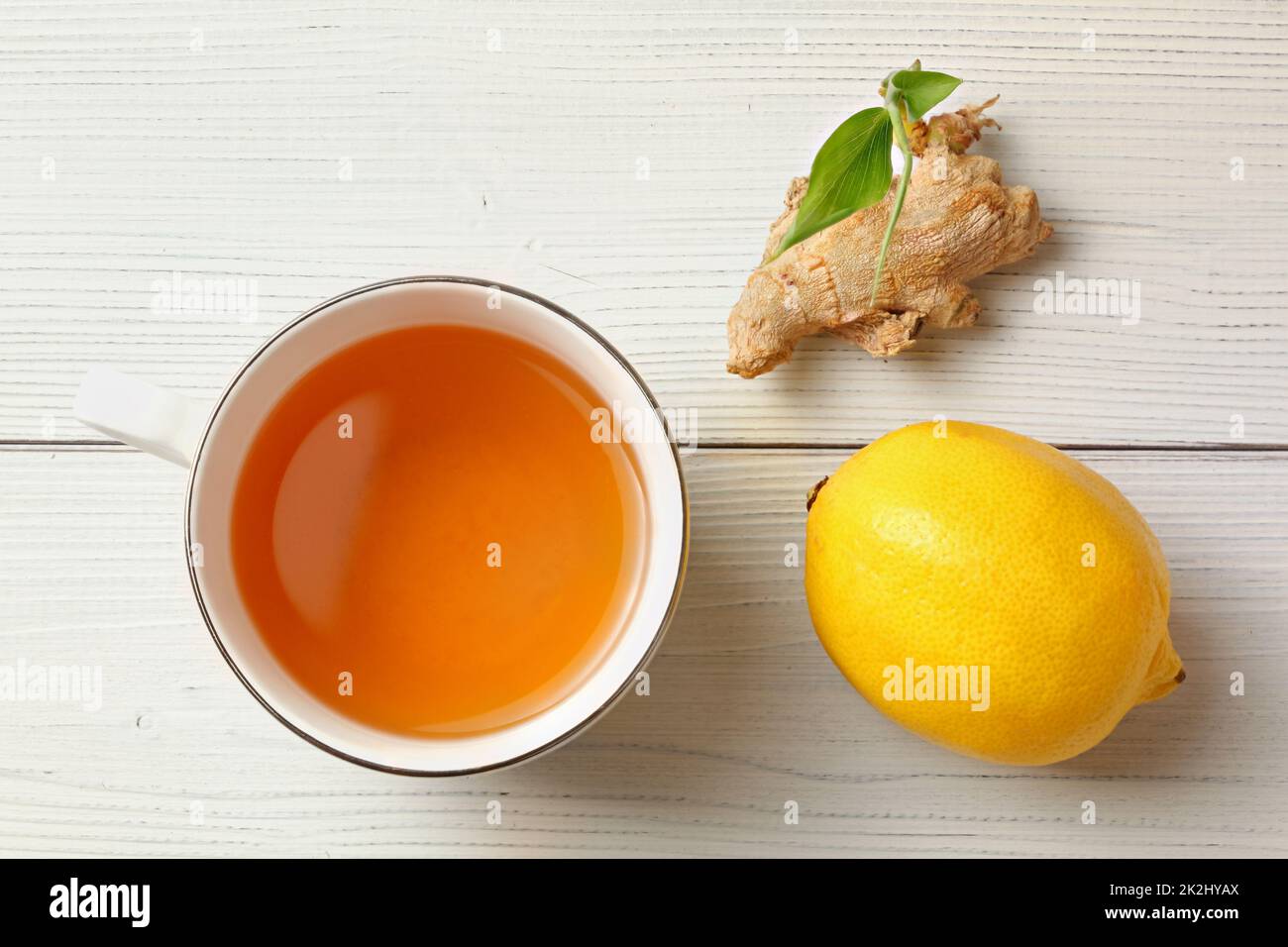 Tabletop view, porcelain cup of freshly brewed tea, dry ginger root with green sprout and whole lemon next to it, placed on white boards desk. Stock Photo