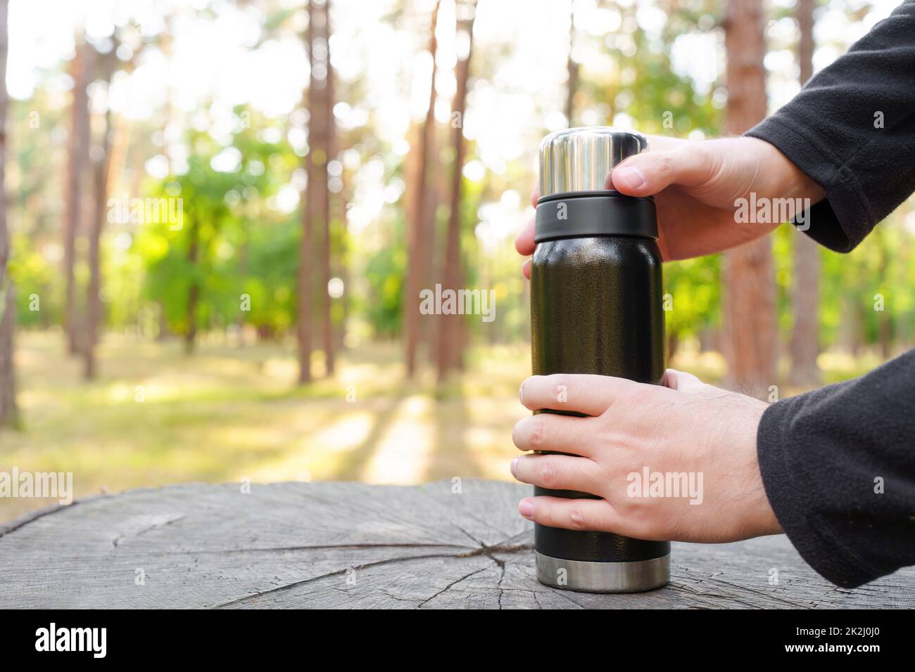 Male hands opening a black thermos bottle using a large tree stump as a coffee table in the forest. Stock Photo