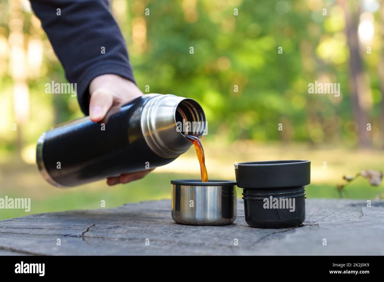 Hand pouring hot coffee from a thermos bottle into a cup placed on a tree stump. Coffee break in the woods. Stock Photo
