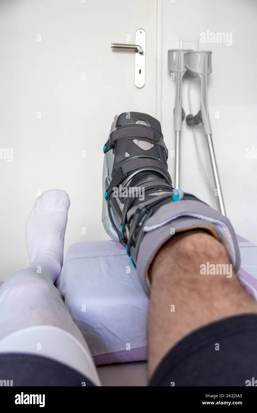 European man after Achilles tendon rupture operation is back home with special physiotherapy shoe and crutches for recovery at home with healthy medicine painkiller drug pills against the hurting leg Stock Photo