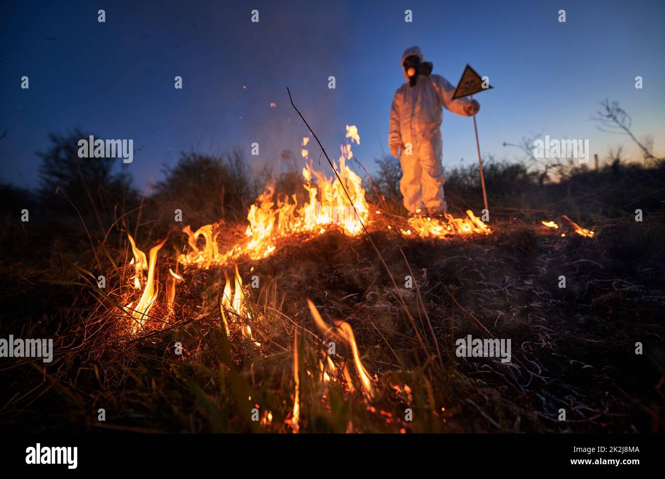 Fireman ecologist working in field with wildfire at night. Man in protective radiation suit and gas mask near burning grass with smoke, holding warning sign. Natural disaster concept. Stock Photo