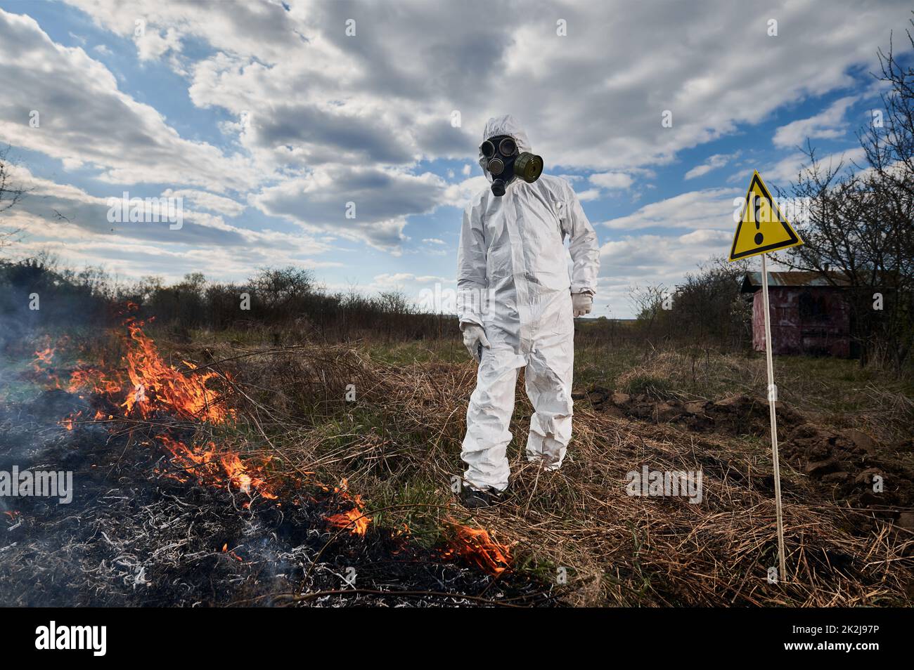 Firefighter ecologist working in field with wildfire. Man in protective radiation suit and gas mask near burning grass with smoke and warning sign with exclamation mark. Natural disaster concept. Stock Photo