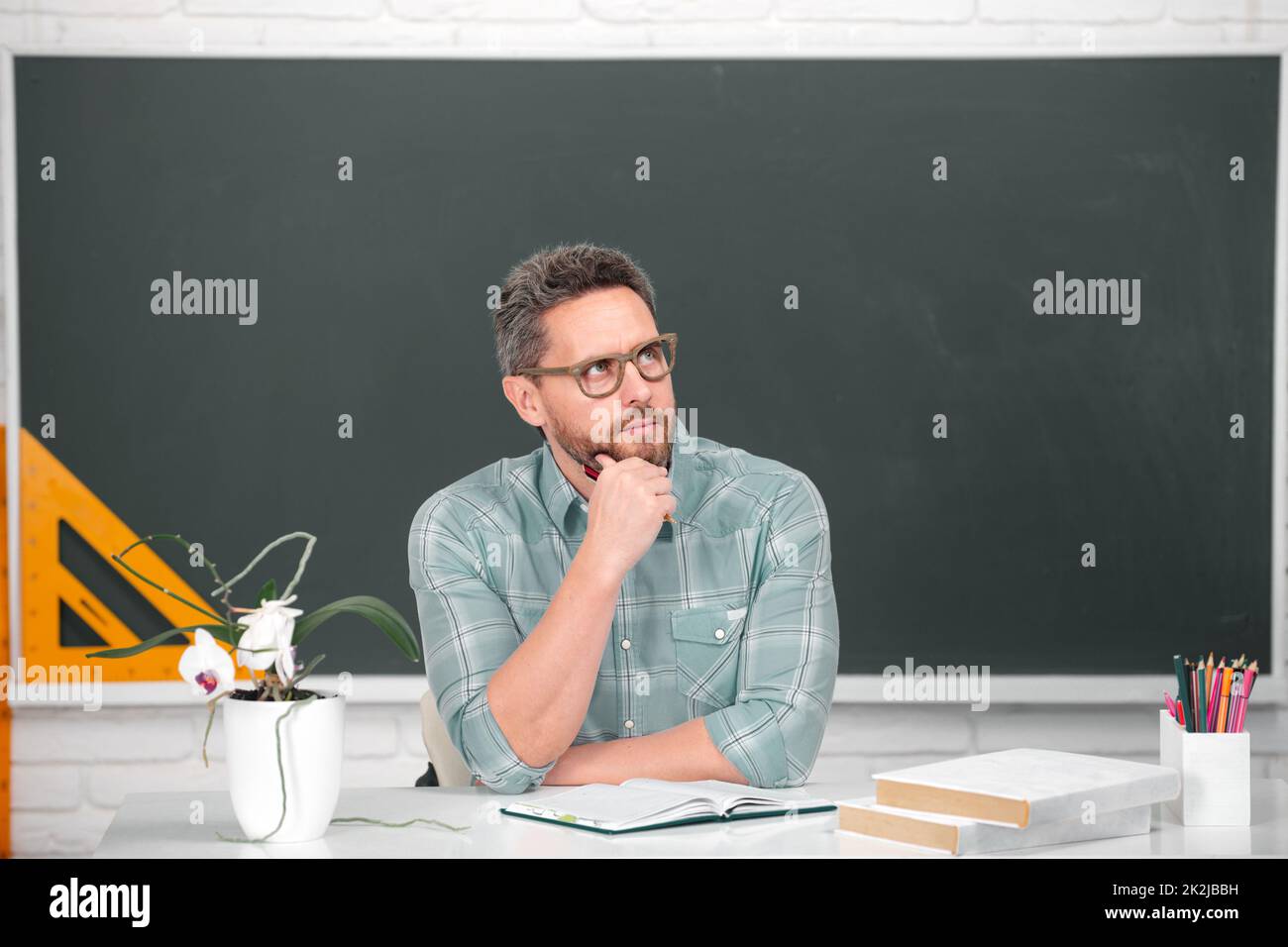 Portrait of confident caucasian male teacher in classroom. Young male thinking teacher with chalk near blackboard in classroom. Stock Photo