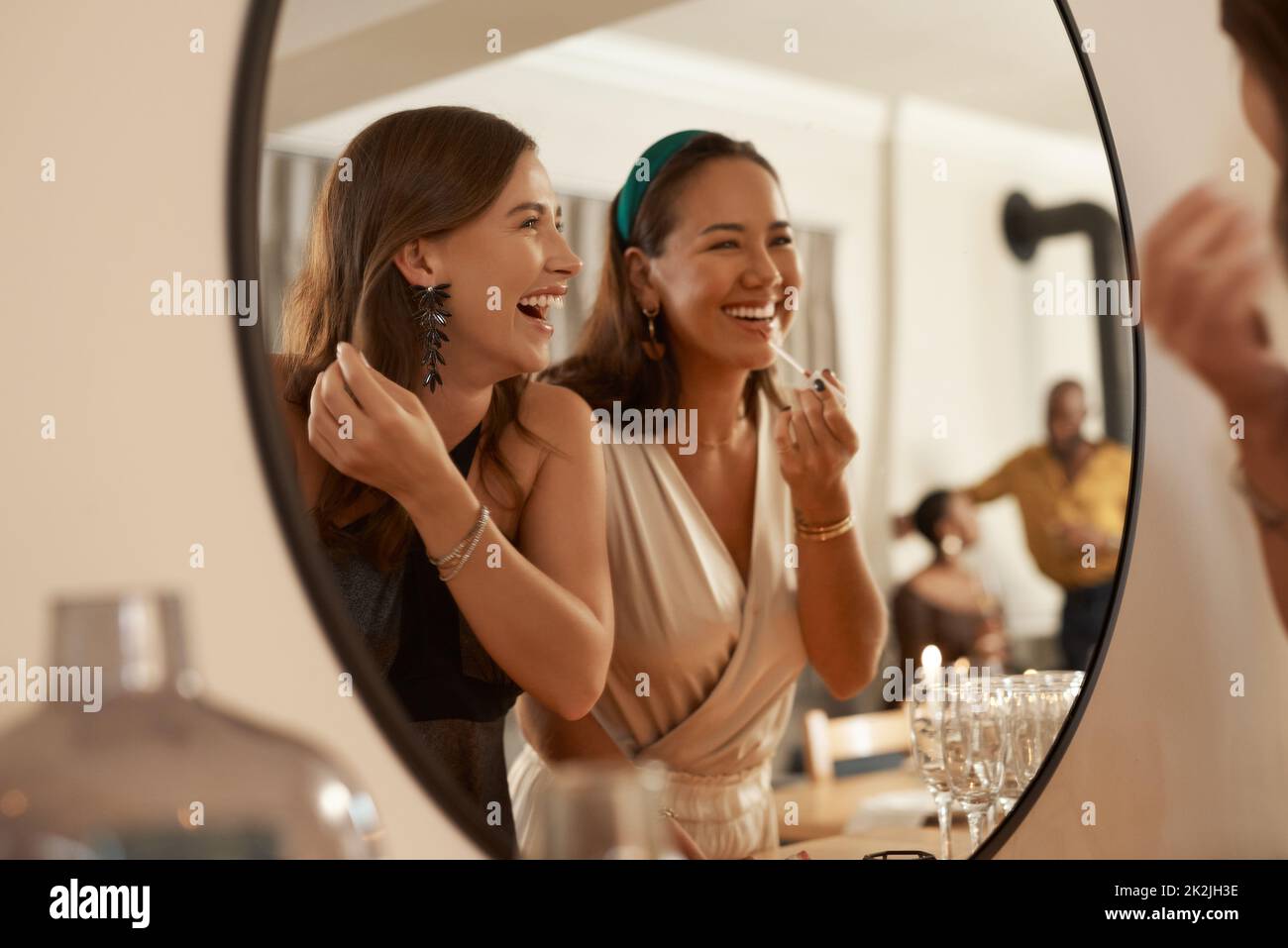 We always have a joke prepared. Shot of two young friends standing together and using a mirror to touch up their makeup at a dinner party. Stock Photo