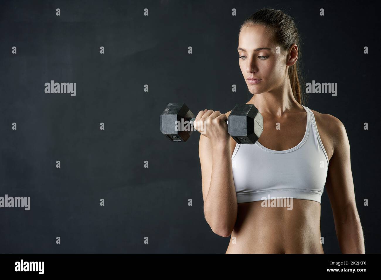 Focusing on her arms. Studio shot of an attractive young woman working out with a dumbbell against a dark background. Stock Photo