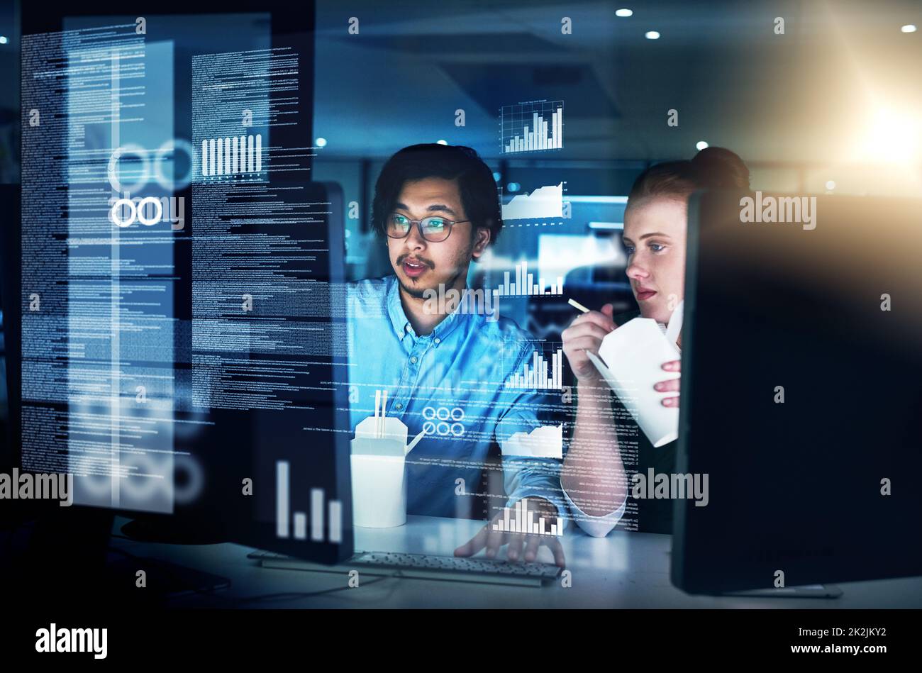 Cracking codes through the night. Shot of two focused young male programmers working together on a computer while eating noodles in the office at night. Stock Photo