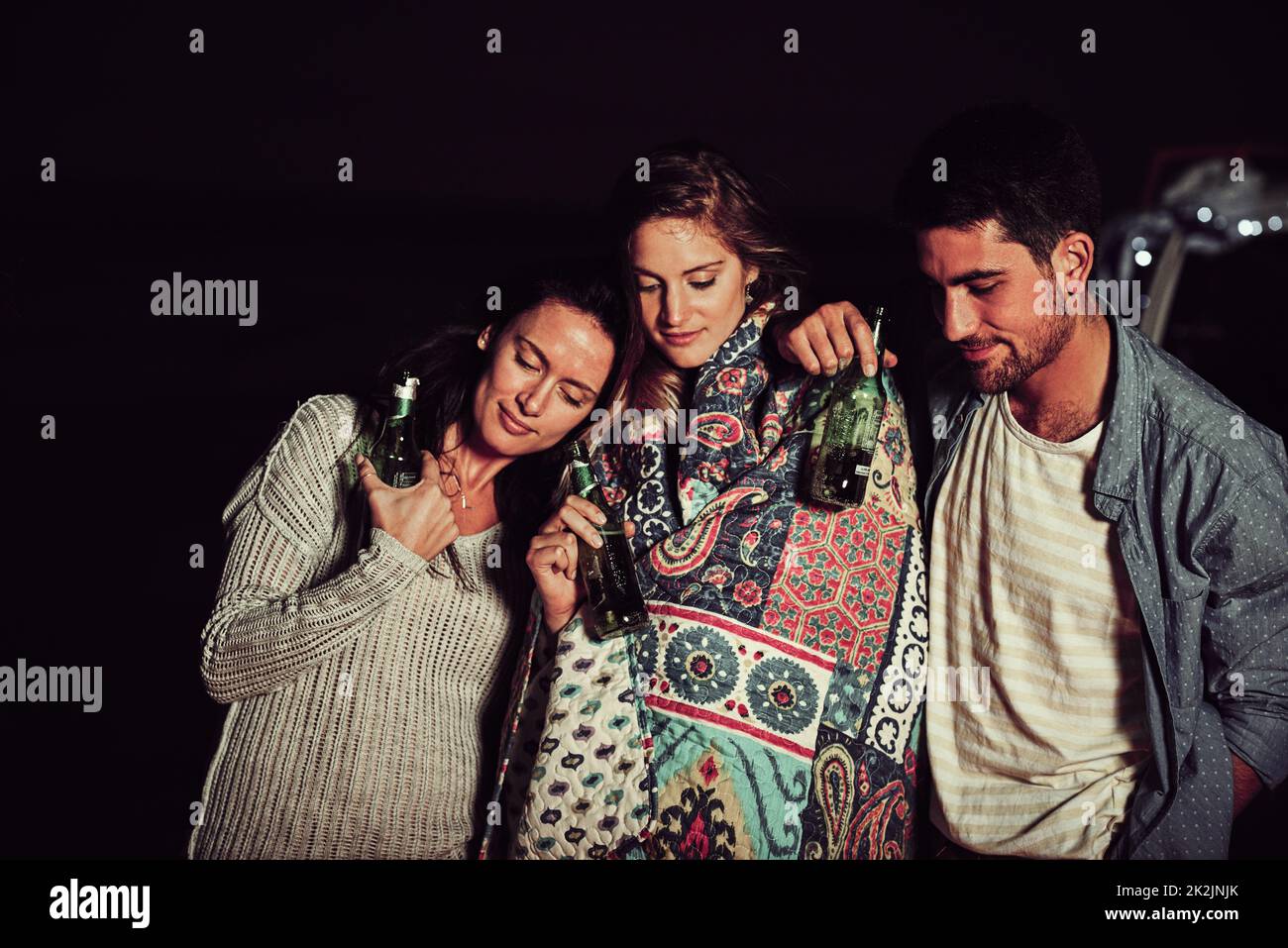 As close as can be. Cropped shot of a group of friends having a drink on the beach at night. Stock Photo