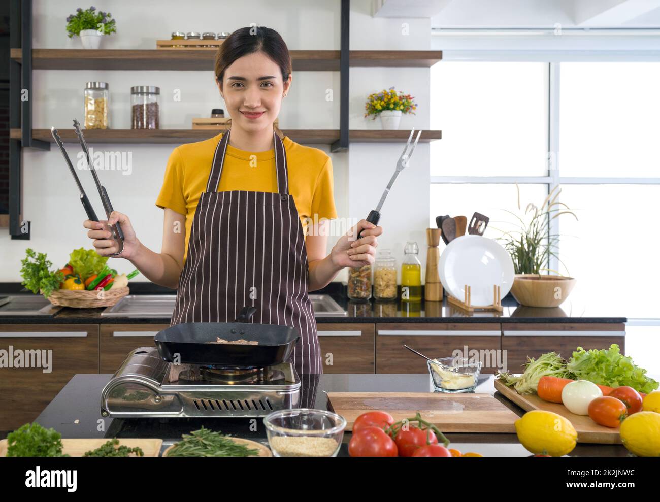 Young housewife holding BBQ Clamp Shovel and Stainless Steel Fork in front of cooking pan. Morning atmosphere in a modern kitchen. The kitchen counter full of various kinds of vegetables. Stock Photo