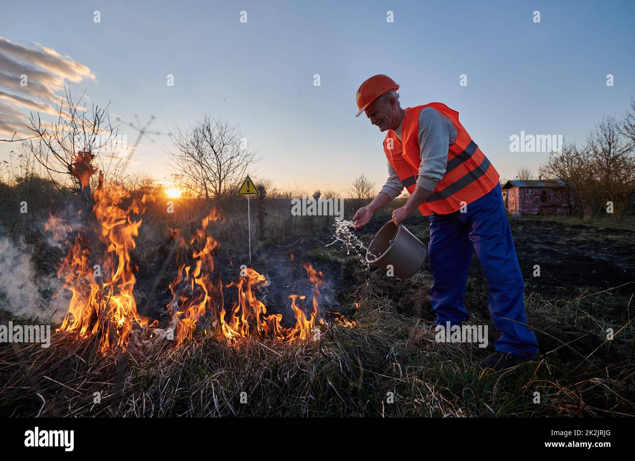 Fireman ecologist extinguishing fire in field with warning sign with exclamation mark end evening sky on background. Male environmentalist holding bucket and pouring water on burning dry grass. Stock Photo