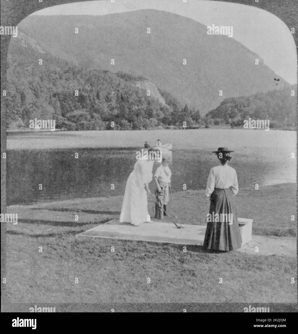 Woman and boy with golf clubs in foreground, 2 people in boat on lake, mountain in background. c1905 Stock Photo