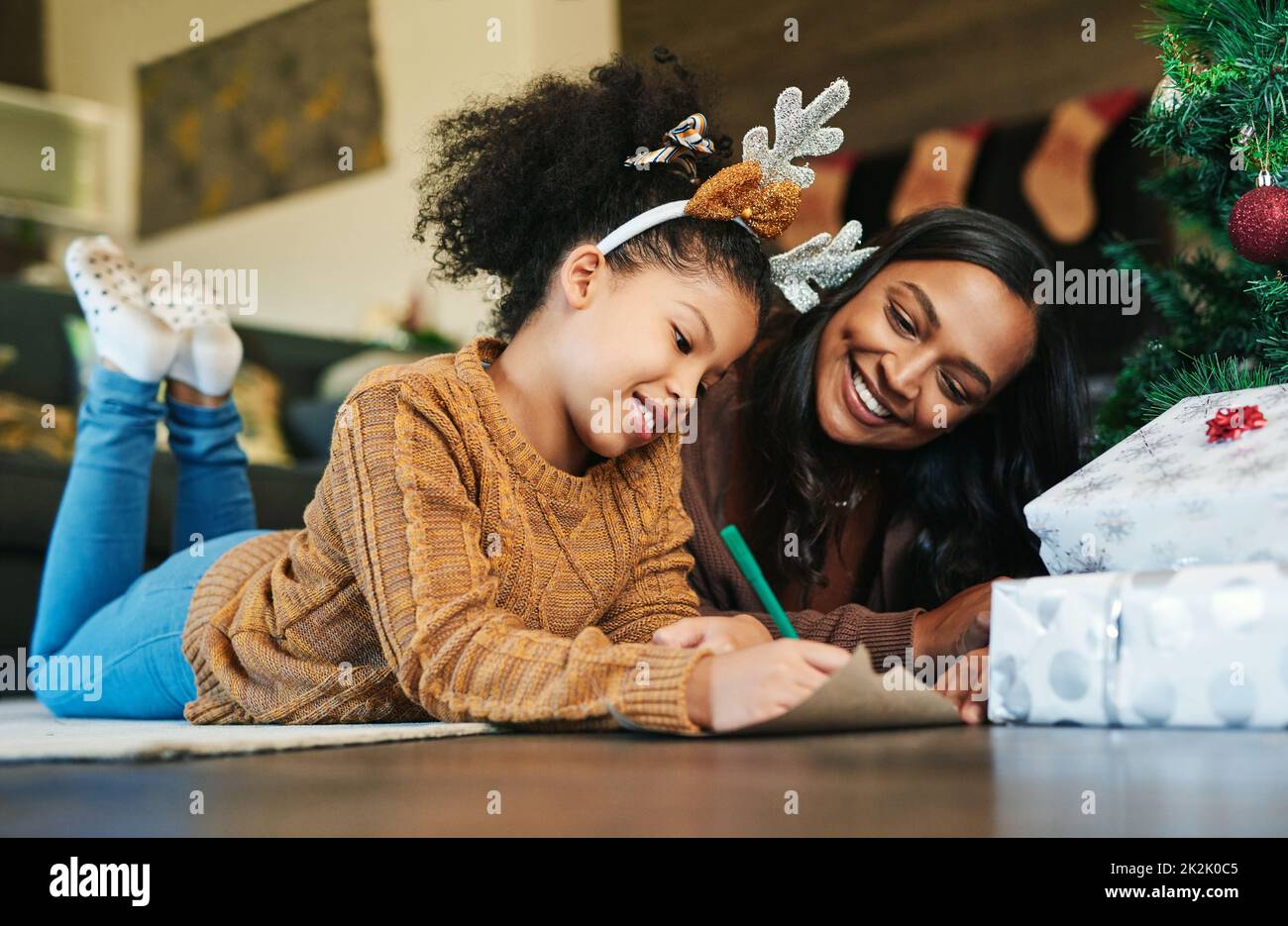 Dear Santa, ask my mom how good Ive been. Shot of a cute little girl writing in a card with her mother during Christmas at home. Stock Photo