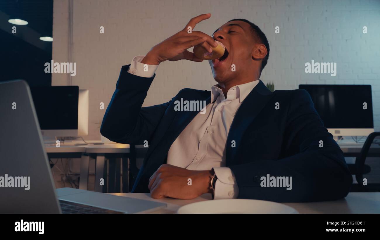 African american businessman eating donut near blurred laptop in office in evening,stock image Stock Photo
