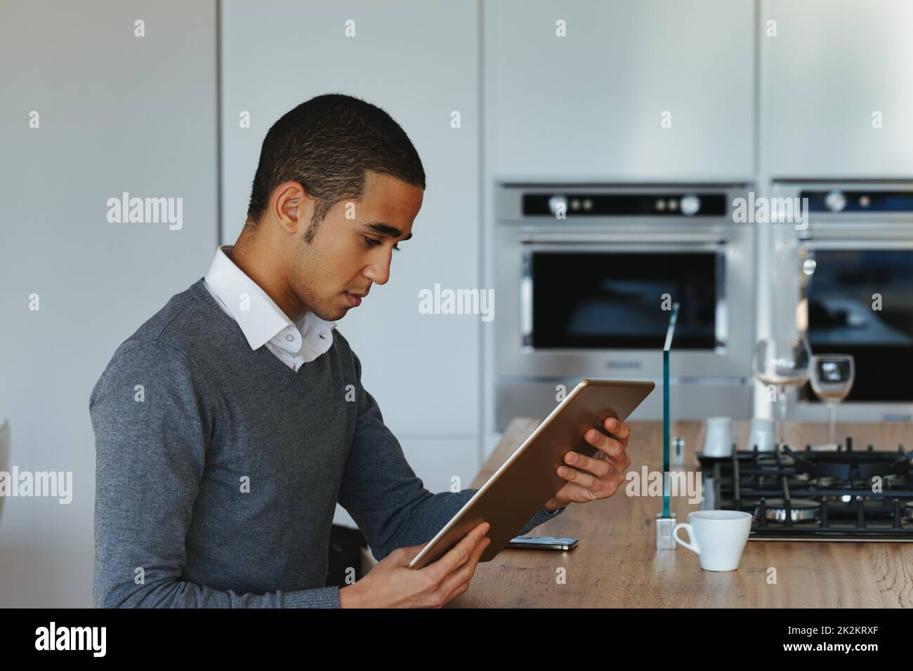 Young Black man seated at a kitchen table reading on a tablet pc Stock Photo