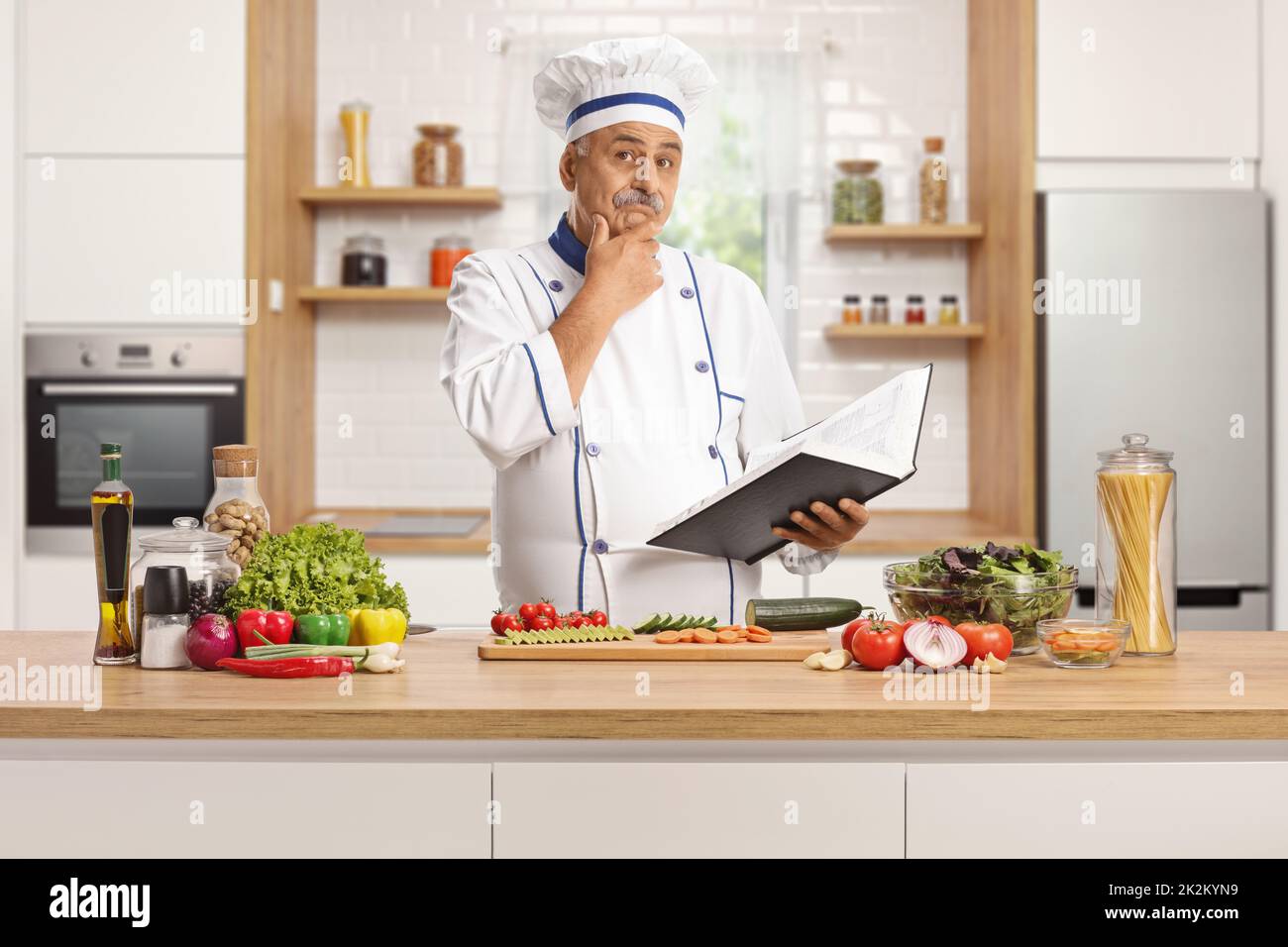 Mature male chef behind a kitchen counter holding a cook book and thinking Stock Photo