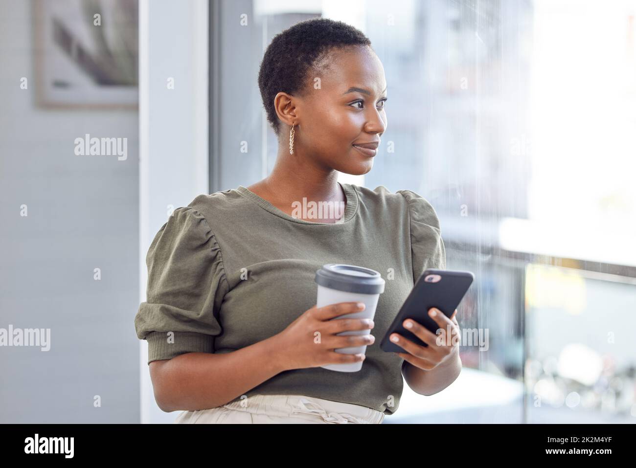 The internet is my link to world out there. Shot of a businesswoman drinking coffee and using her cellphone while looking out her office window. Stock Photo