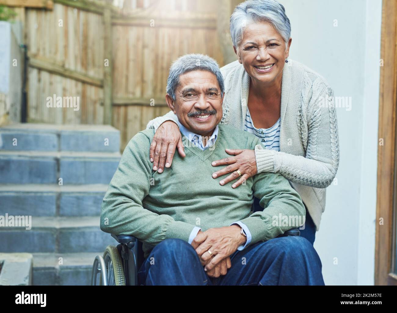 Shes means the world to me. Portrait of a cheerful elderly man seated in a wheelchair while being held by his loving wife outside at home during the day. Stock Photo