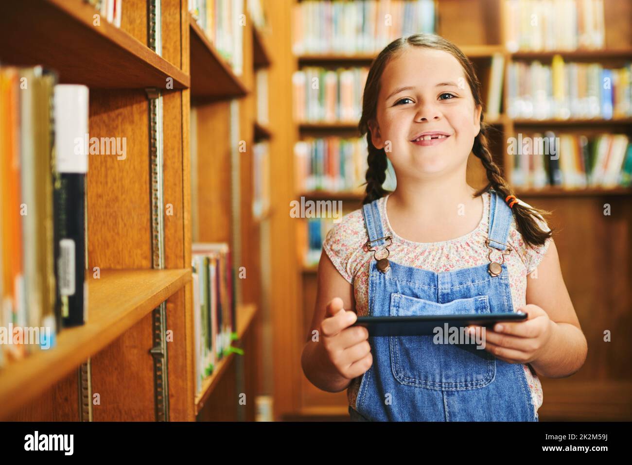 This is almost like a book. Portrait of a cheerful young girl browsing on a digital tablet while standing inside of a library during the day. Stock Photo