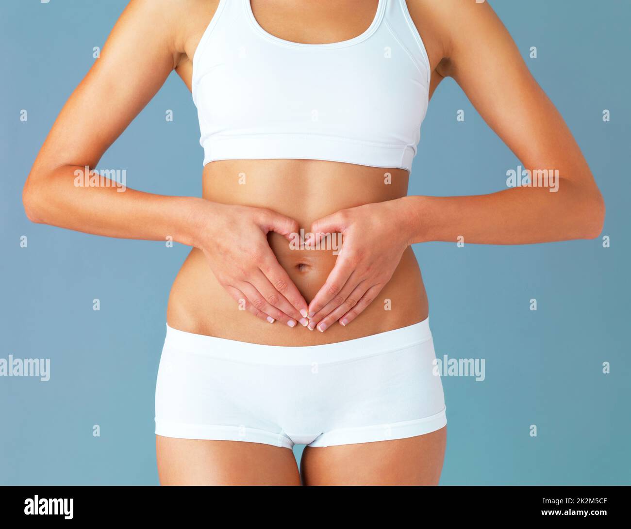 For the greatest wealth, invest in your health. Cropped studio shot of a fit young woman making a heart shaped gesture over her stomach against a blue background. Stock Photo