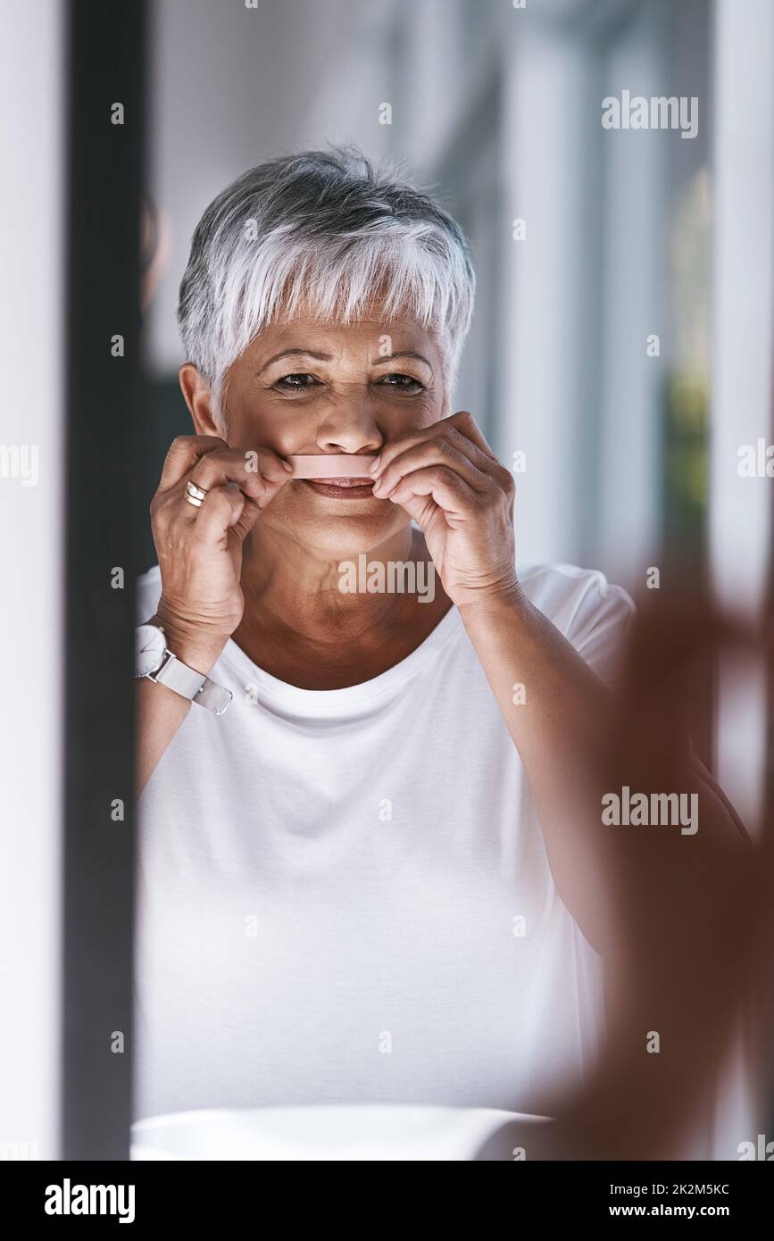 She makes sure she is looking flawless everyday. Portrait of a cheerful mature woman waxing her moustache while looking into a mirror at home. Stock Photo