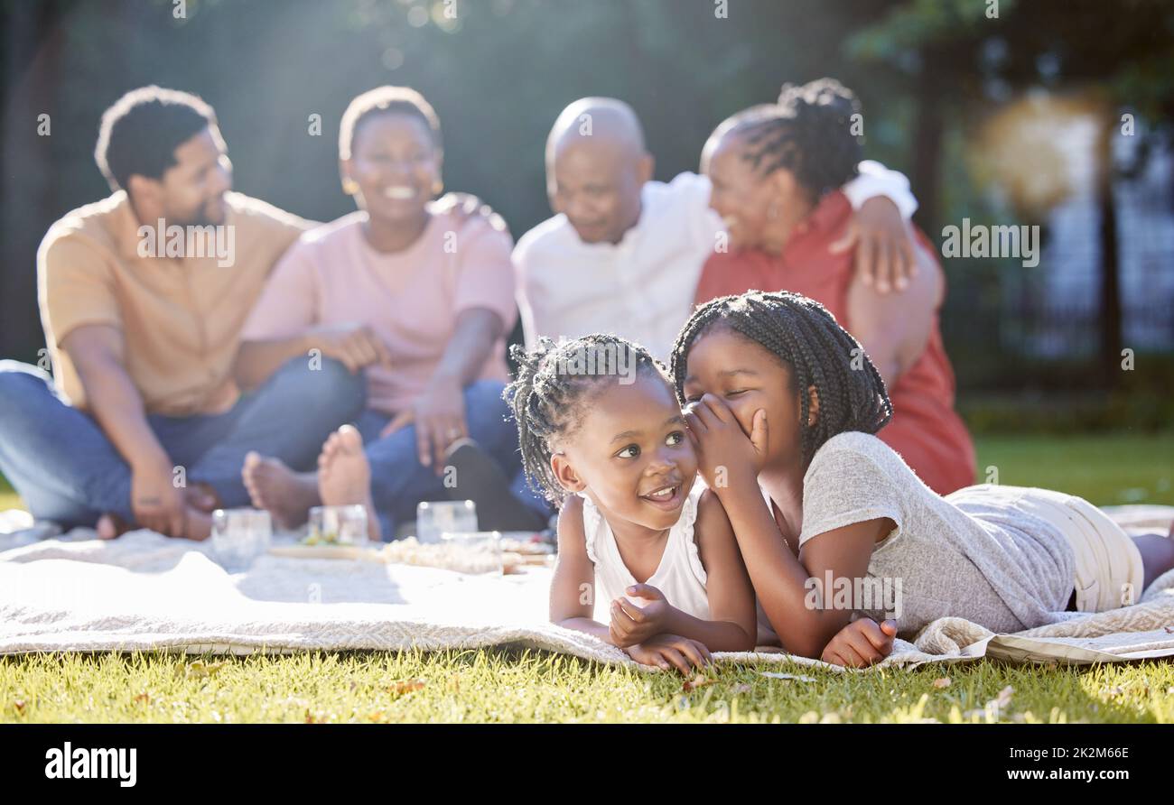 Secret, sister and children with a girl whispering to her sibling and a black family in the background. Kids, mystery and gossip with a female child Stock Photo