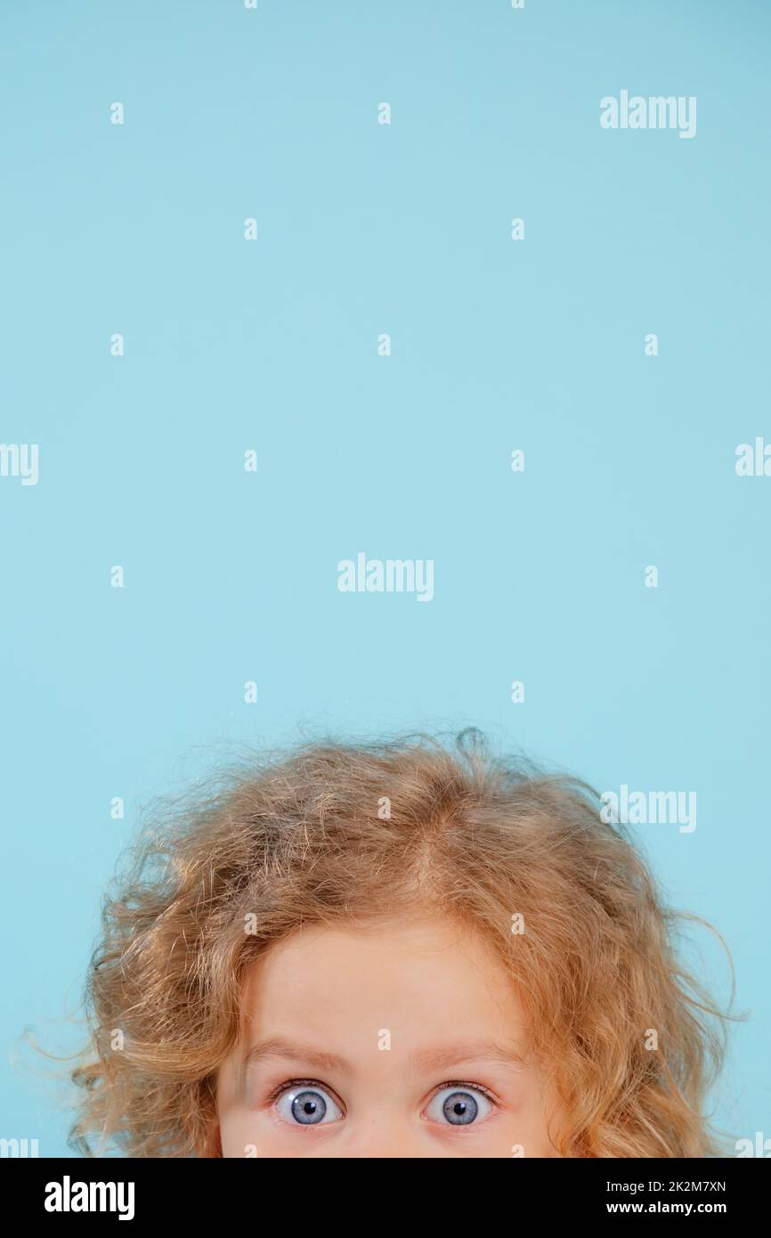 Cropped photo of amazing blue-eyed little girl with curly fair hair looking surprisingly shocked on blue background. Stock Photo