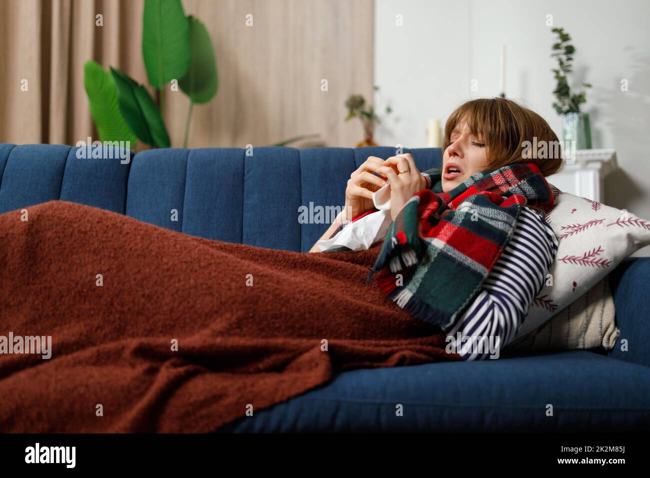 Depressed woman with a runny nose lying under a blanket on the sofa in