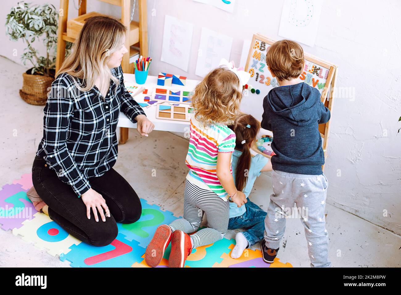 Little kids and educator studying numbers and alphabet on magnetic board sitting on floor in playroom backview. Interesting lesson for kindergartners Stock Photo