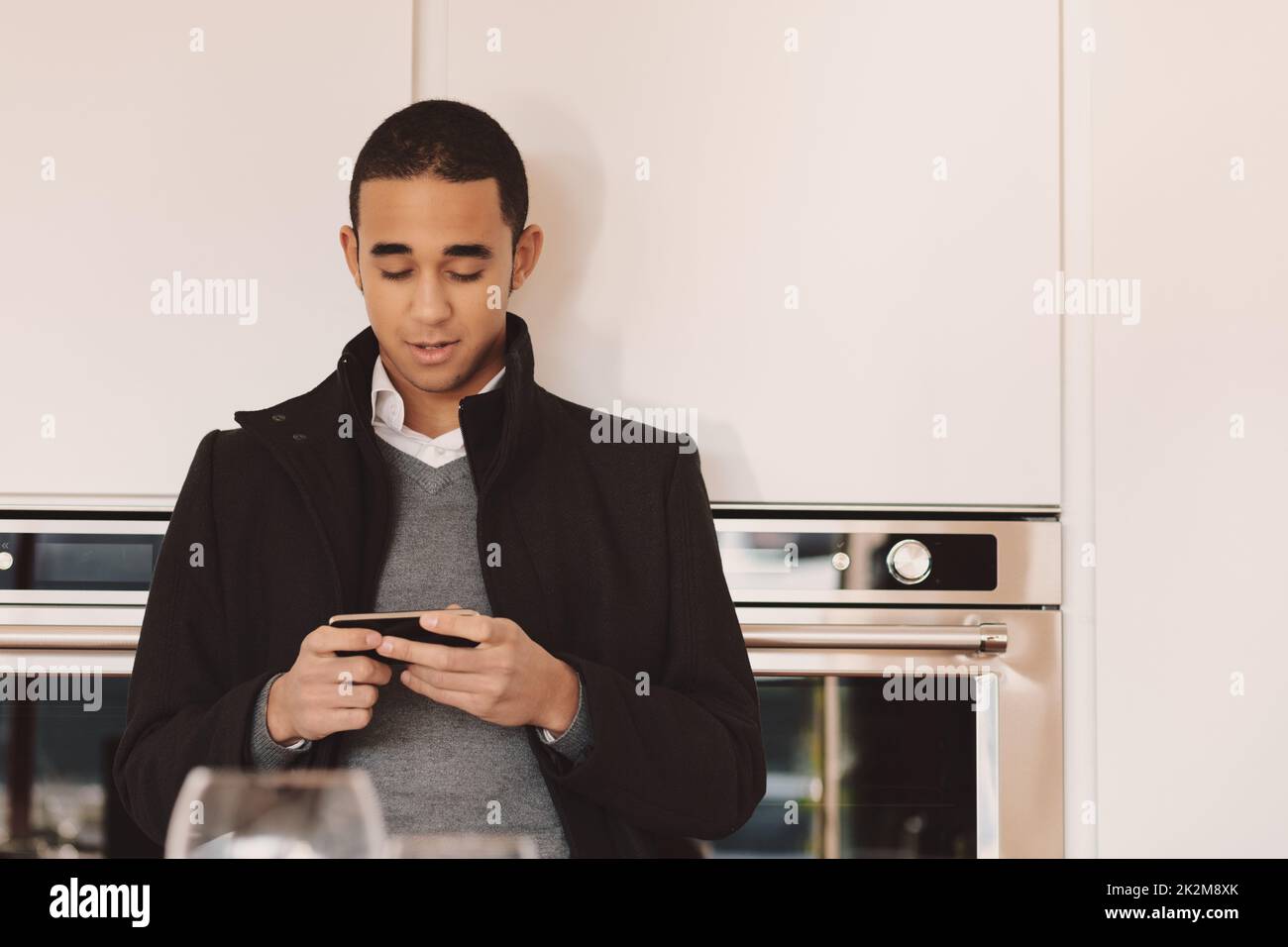 Young man in overcoat standing in a kitchen checking his mobile Stock Photo