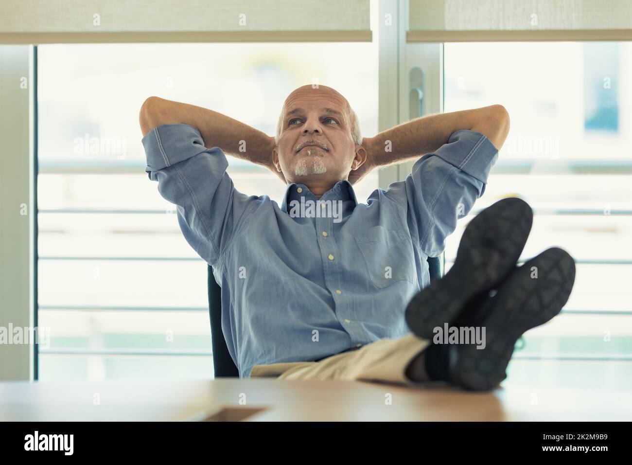 Man in office relaxes with arms crossed behind his head and feet on