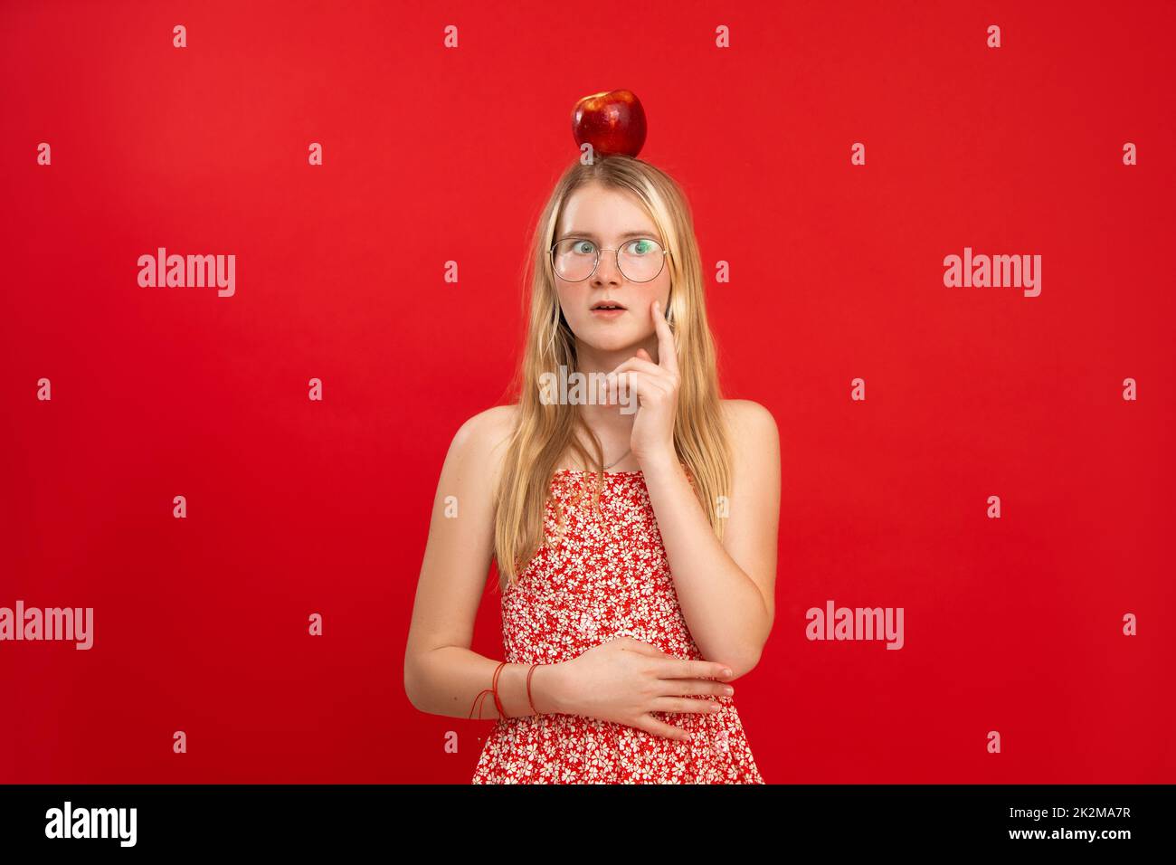 Portrait of puzzled teenage girl with fair hair, big red apple on head wearing glasses touching cheek with index finger. Stock Photo