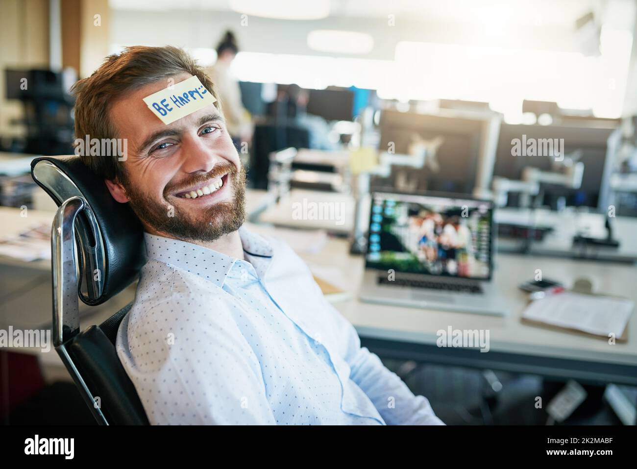 He is one happy camper. Shot of a cheerful young businessman with a sticky note on his forehead saying be happy while being seated in the office. Stock Photo