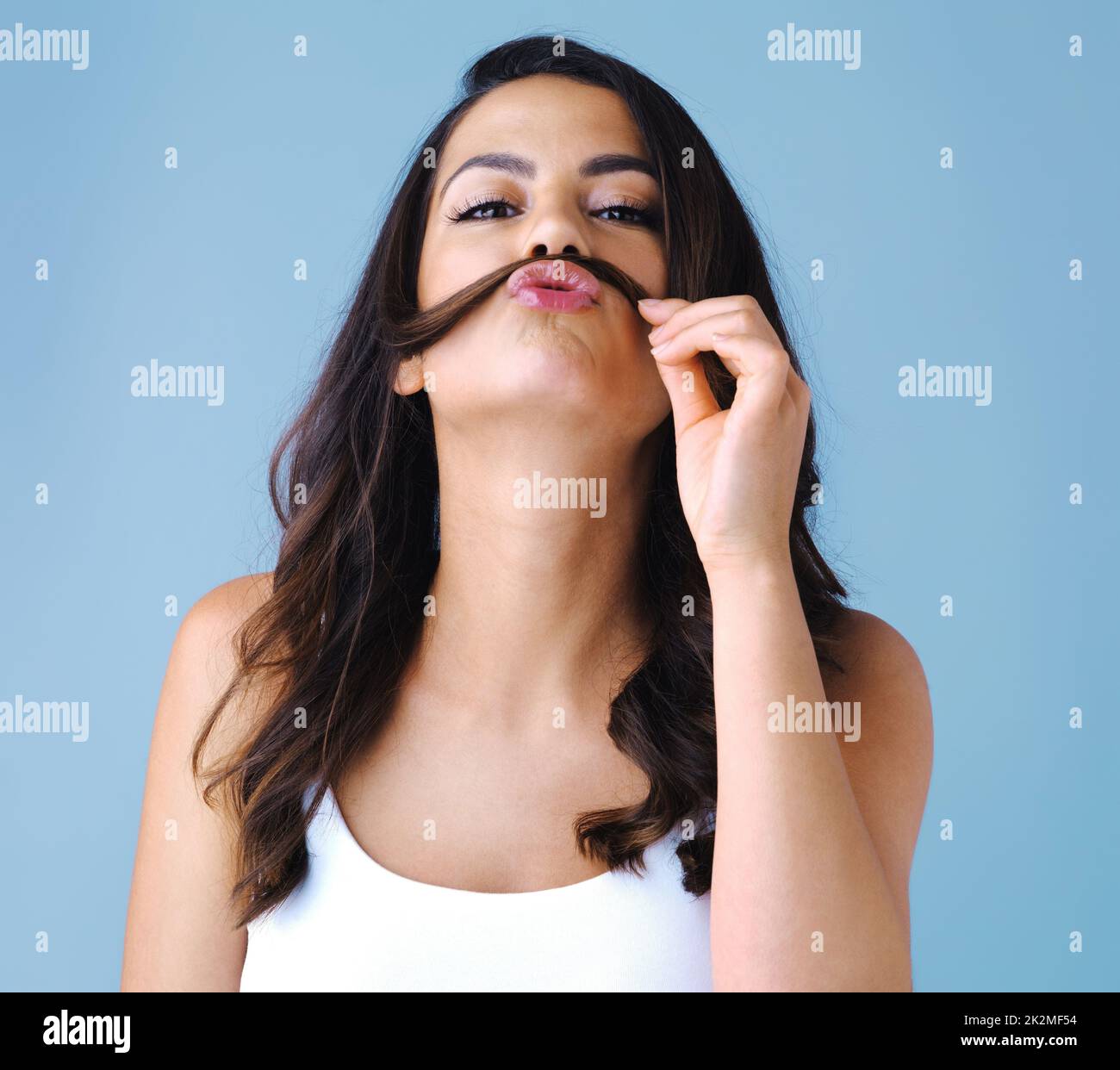 Its all part of her charm. Studio shot of an attractive young woman playfully making a mustache with her hair against a blue background. Stock Photo