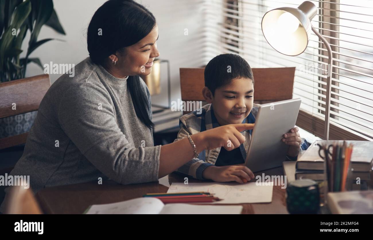 Teaching with tech gets them ready for the real world. Shot of an adorable little boy using a digital tablet while completing a school assignment with his mother at home. Stock Photo